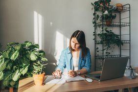 Woman using a calculator to calculate finances at a desk surrounded by greenery and natural sunlight.