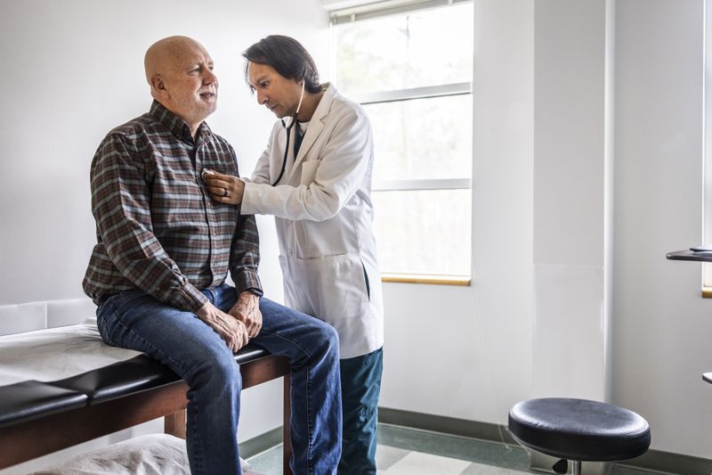 A man sits on a table in a doctor's office while the doctor uses a stethoscope to listen to the man's heart