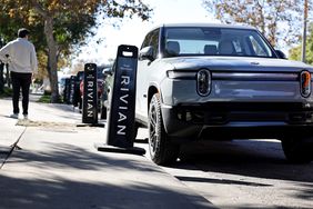 A person stands on the sidewalk to the left of several Rivian electric vehicles parked along the street.