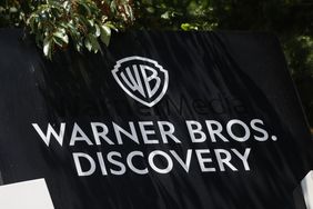 The white Warner Bros. Discovery logo is seen on a black sign, with leaves above the sign.