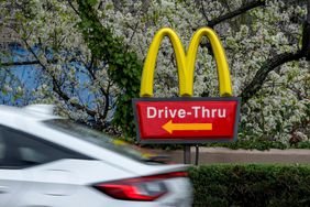 The "Golden Arches" are seen on a McDonald's Drive-Thru sign, with a white car driving by and white and green plants in the background.