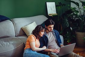 A man and a woman sit on the floor against a couch looking at a laptop