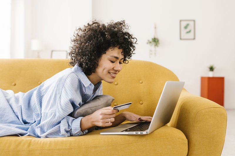 A woman uses a laptop with a credit card on the couch.