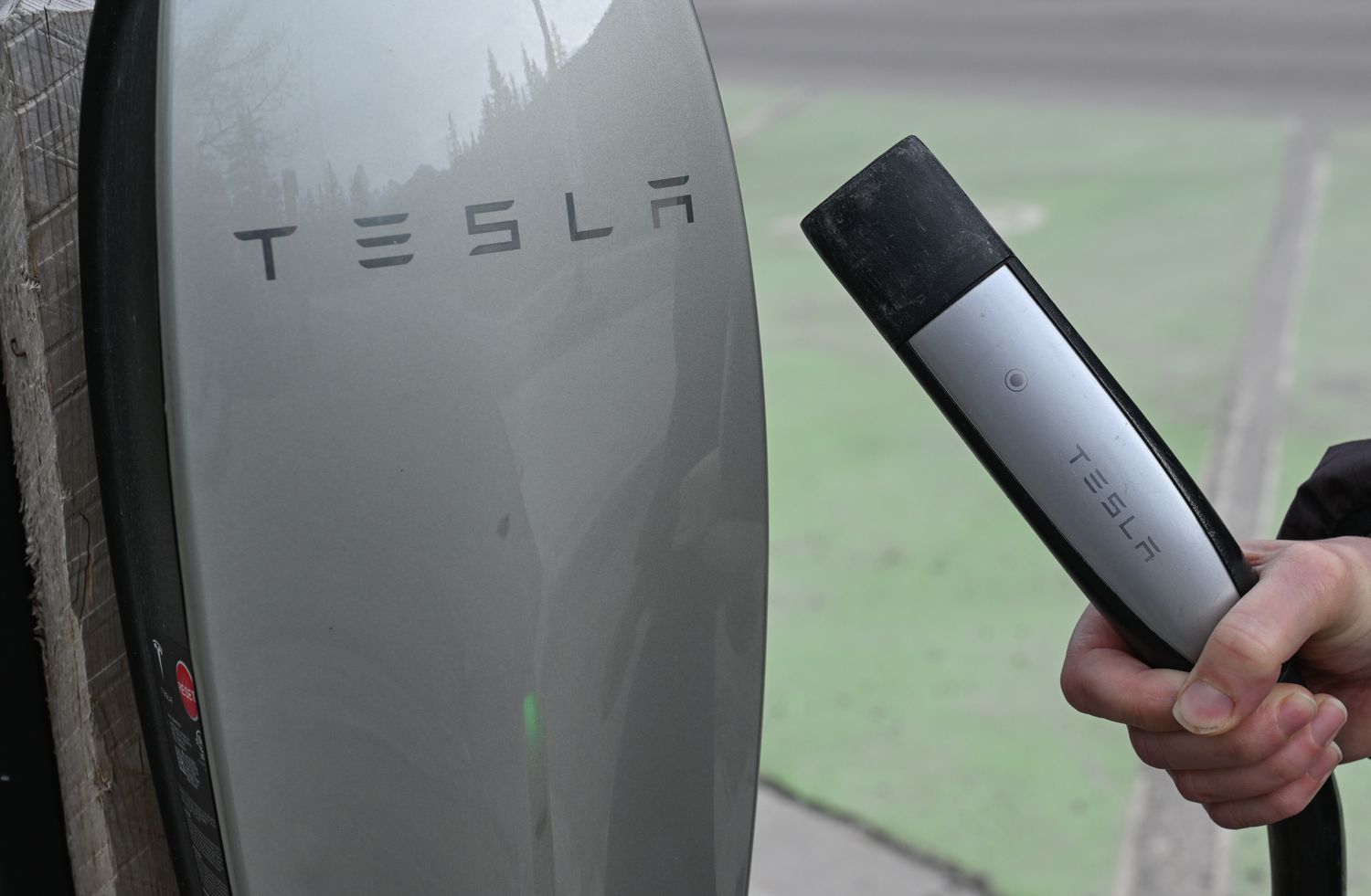 The Tesla logo is seen on the side of a charging station, with a hand holding a charging cable next to the station.
