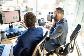Two men sitting at a desk with their back to the camera, discussing data on a computer monitor.