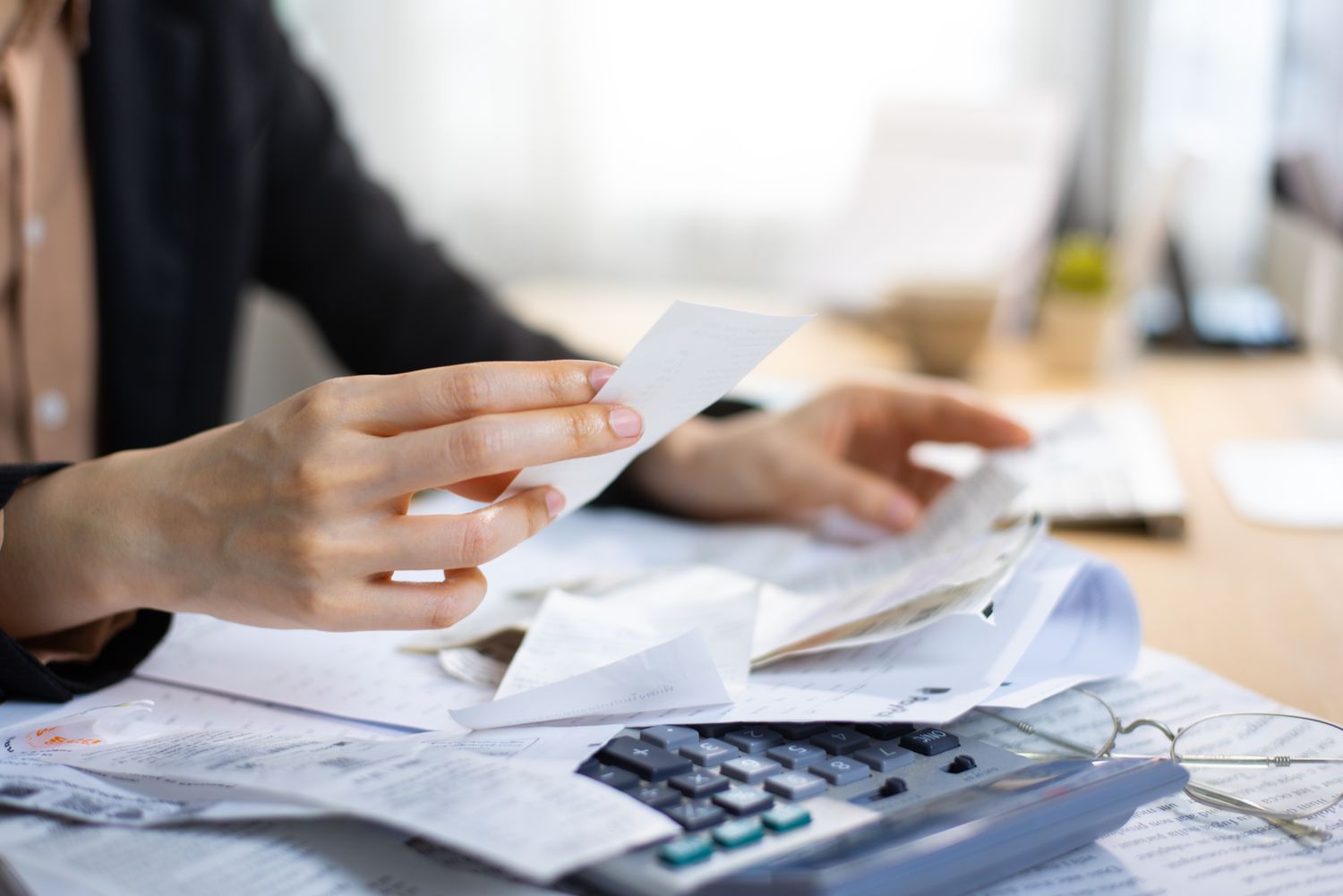 Hands holding receipts with more receipts, a calculator and glasses on the desk