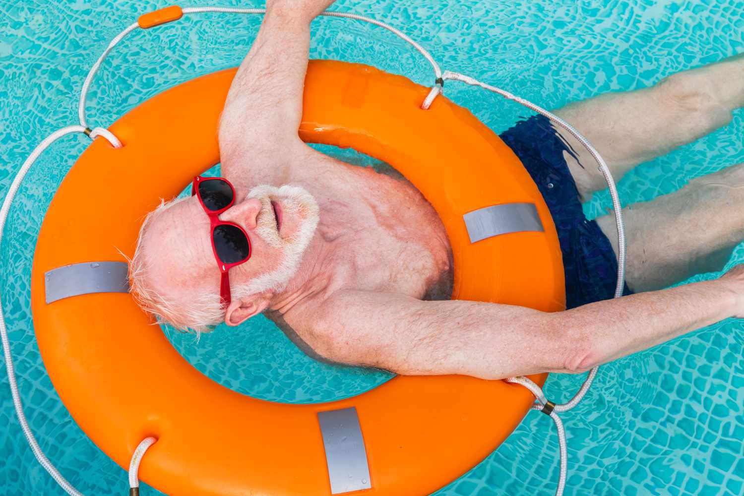 Older man wearing sunglasses in a swimming pool using a life preserver to float. 