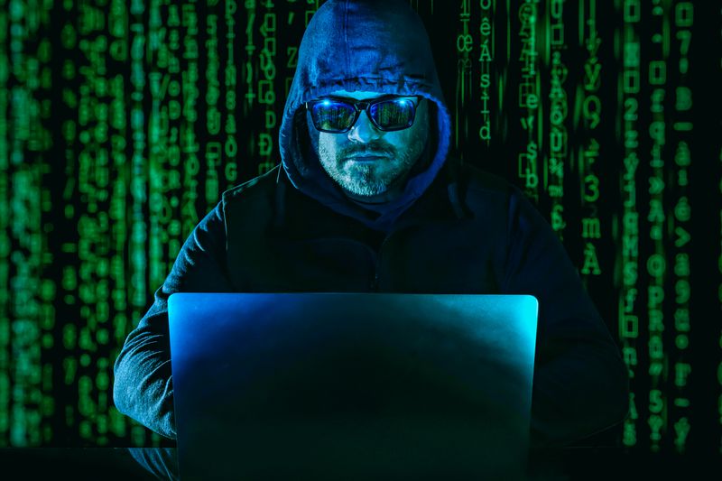 An anonymous person wearing a hooded jacket and sunglasses uses a laptop in front of scrolling rows of cryptocurrency symbols