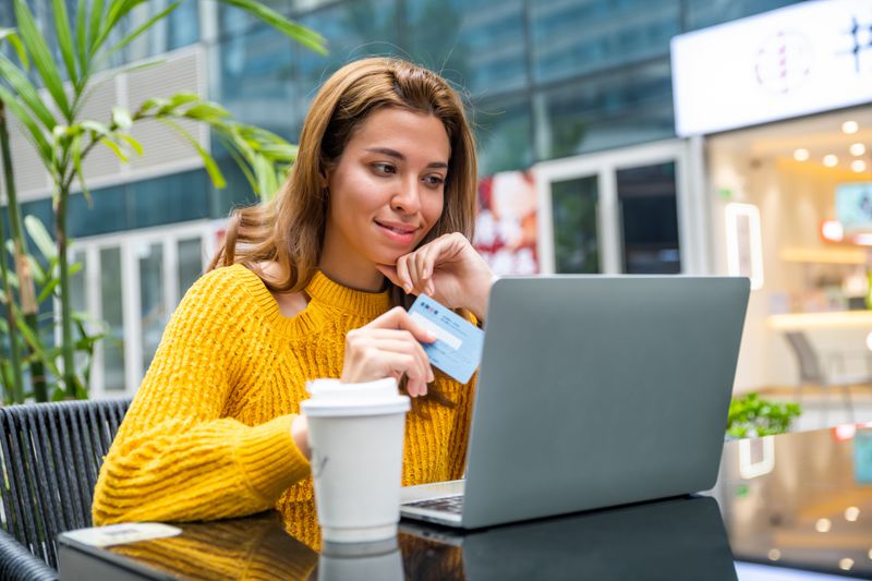 Young woman uses credit card while looking at her laptop at a table outside, with a thoughtful look on her face.