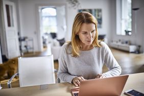 A woman sits at a counter in an apartment with a laptop and smartphone.