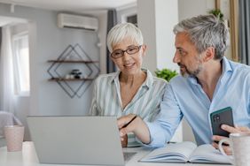 A man and a woman seated together at a table with a laptop, document, and smartphone, engaging in discussion