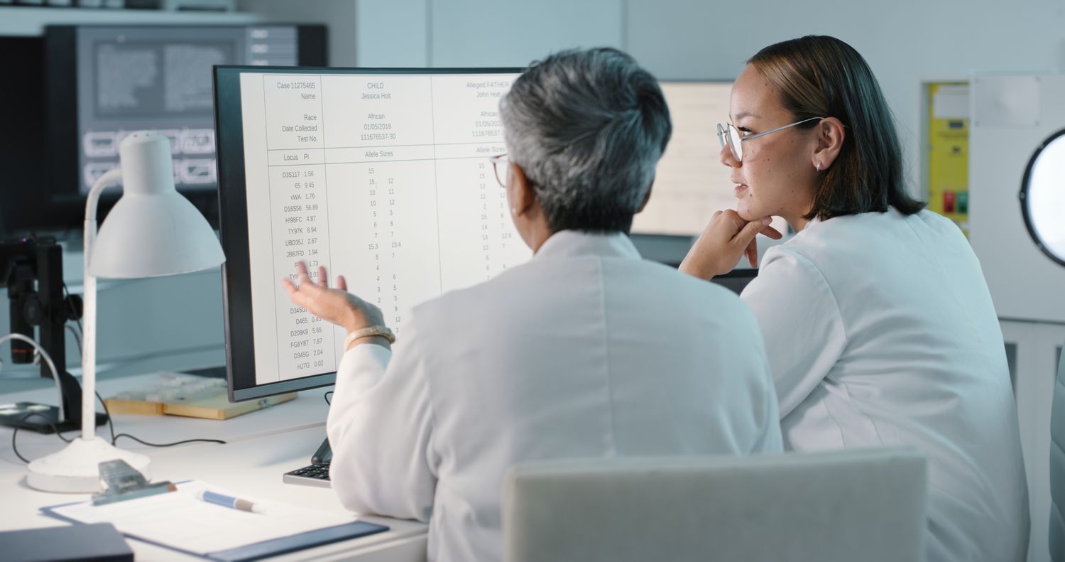 Two women wearing lab coats look at data on a computer screen in a hospital setting