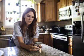 A woman stands at a kitchen counter looking at her smartphone