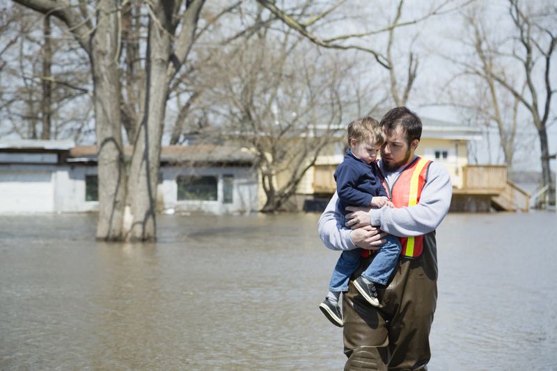 A father and son stand in a flooded street in front of a partially submerged suburban home.