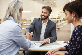 A real estate agent makes a sale, sitting at a table and happily shaking hands with a client.