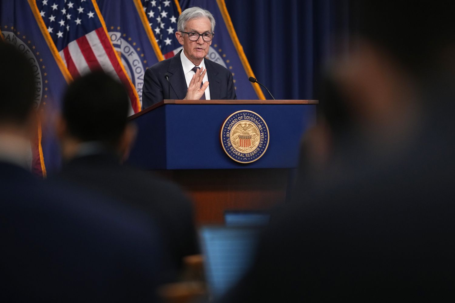 Jerome Powell stand in front of U.S. flags and Federal Reserve flags. He's in front of a podium, facing a crowd of reporters who are out of focus.