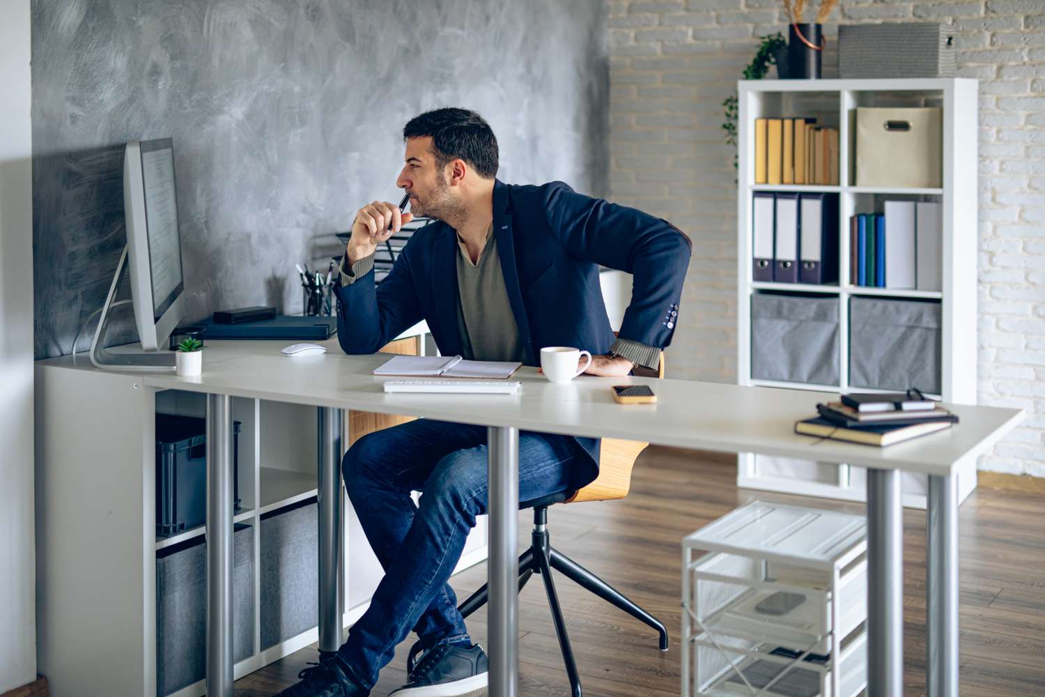 Man sitting at a desk looking at a computer monitor while holding a pen to his mouth.