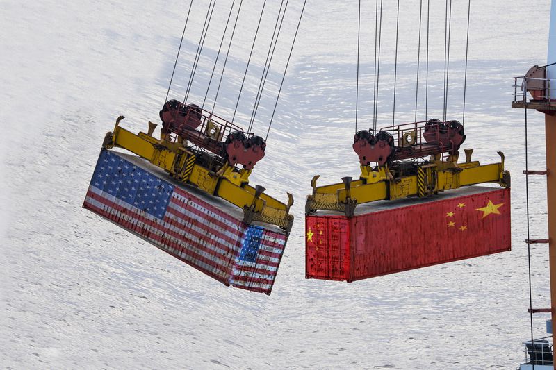 Two containers being moved over water. One is wrapped in the colors of the American flag. The other wrapped in the Chinese flag. 