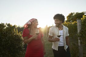 Two people holding wine glasses walking through a vineyard, smiling and enjoying the setting
