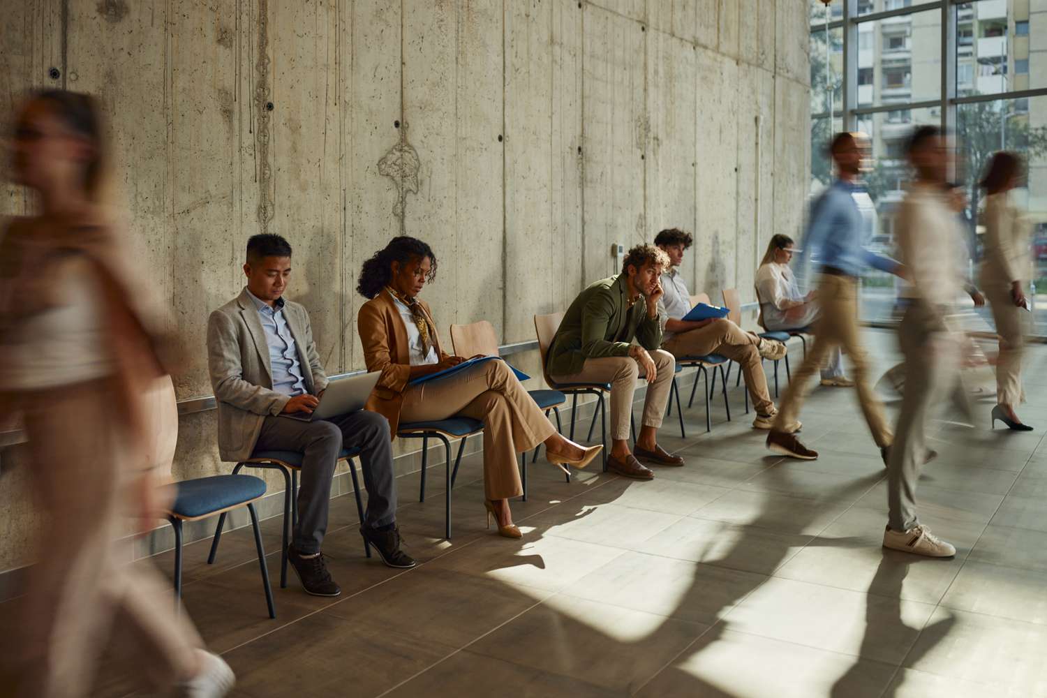 A group of despondent-looking applicants wait in a row of hard chairs lined against a wall waiting on an job inverview.