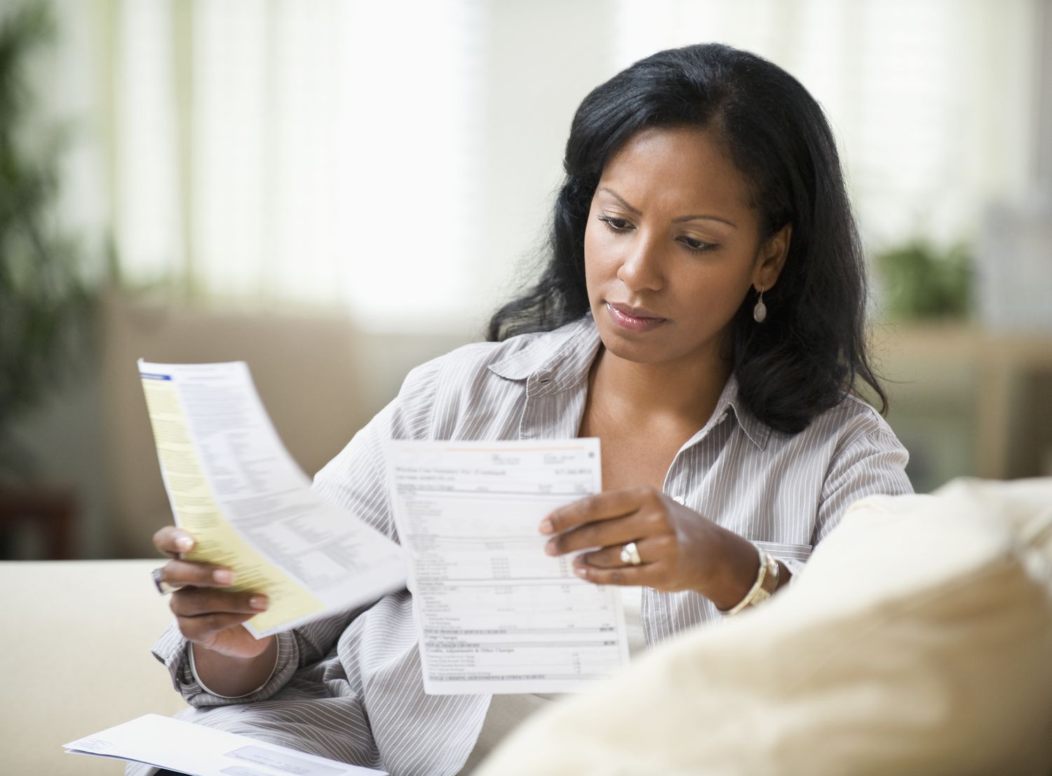 A woman sits on a sofa holding a bill in each hand, looking at them seriously.