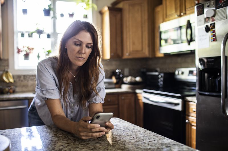 A woman stands at a kitchen counter looking at her smartphone
