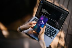 A person holds a smartphone with a robot logo on the screen, while working on a laptop