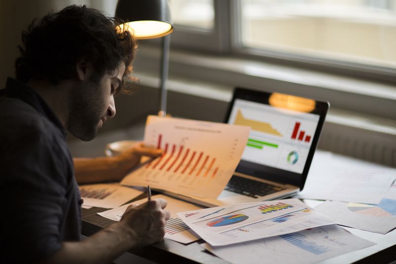 Person writing with a paper with bar graphs in hand and a computer with charts nearby.