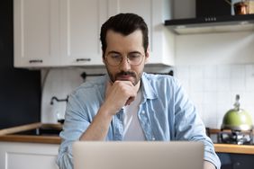 Man in his 30s sitting in his kitchen and pondering something on his laptop