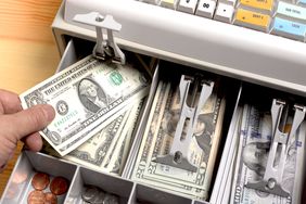 close up shot of the open drawer of a cash till, full of American dollars, with a person putting a $1 bill in.