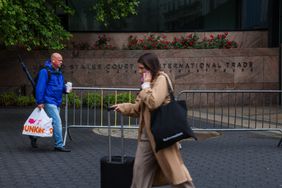 Two individuals walking near the United States Court International Trade building, one carrying a bag and coffee, the other pulling a suitcase