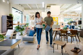 A couple shopping for furniture in a modern showroom, examining home decor items such as a patterned basket. The space features contemporary furnishings, greenery, and large windows allowing natural light to fill the room.
