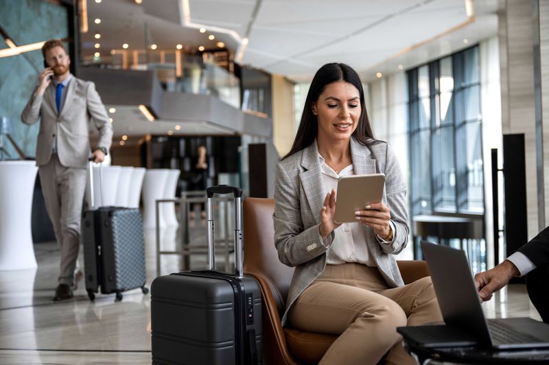Businesswoman on tablet in hotel lobby with suitcase and other traveling workers around her