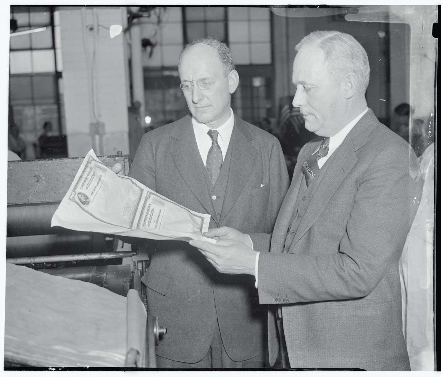 Secretary of the Treasury Henry Morgenthau Jr., inspects the first $100 bond handed to him by Alvin W. Hall, director of the Bureau of Engraving and Printing.