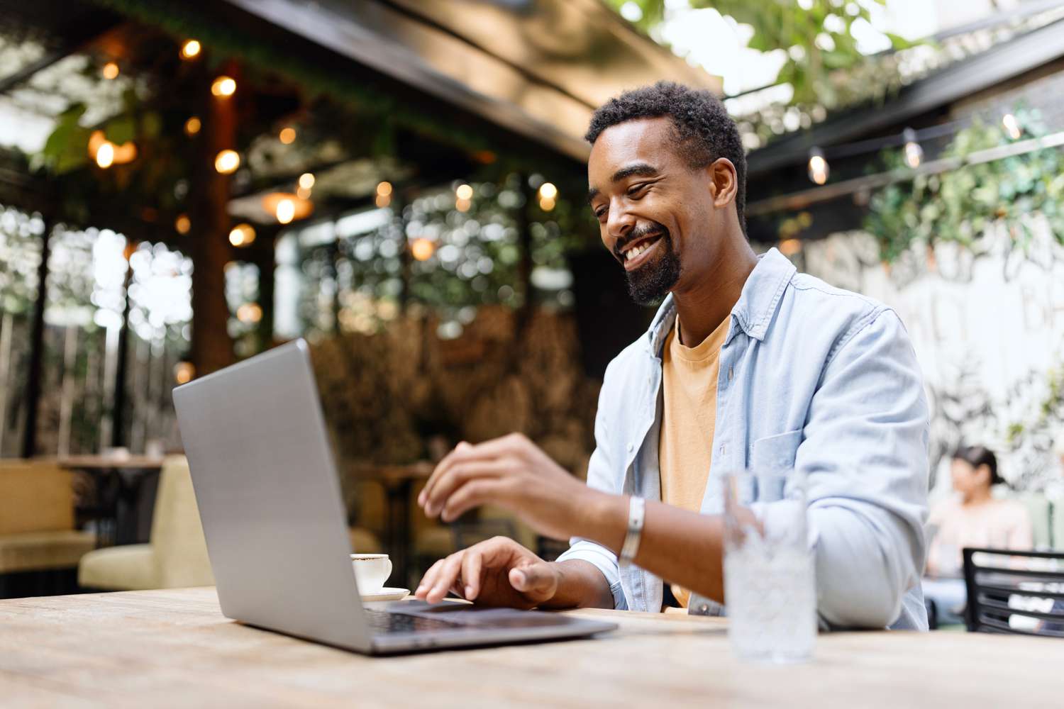A happy man sits in a cafe using a laptop with a big smile on his face.
