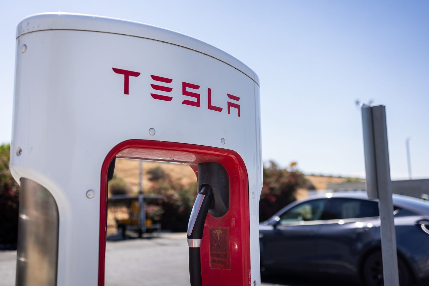 The Tesla logo is seen on a charging station, with a car in the background.