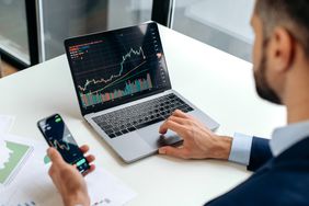 Trader checking market index on laptop while making trading decision on the phone.