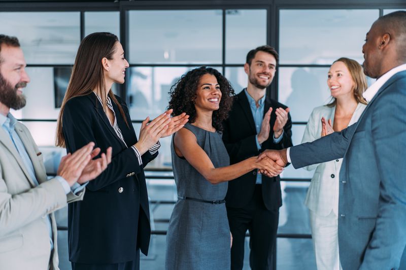 A group of employees applaud as two business owners shake hands after completing a business agreement between the two companies.