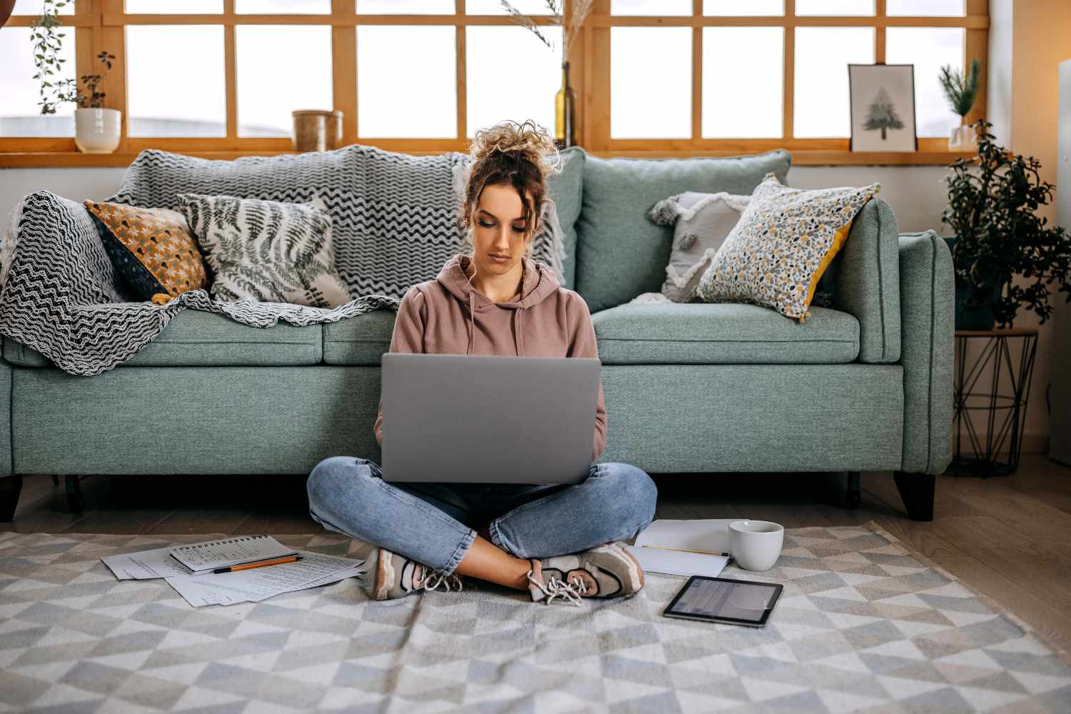 A woman sits on the floor in front of a green couch; she types on a laptop with papers, a tablet, and a mug on the floor around her