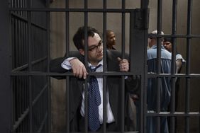 A person wearing a suit stands holding the bars in a prison cell, with other individuals in the background of the cellblock