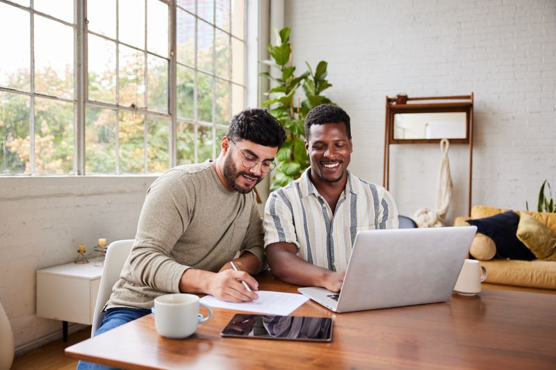 Young multiracial gay couple smiling while going over their home finances together at a table in their living room at home