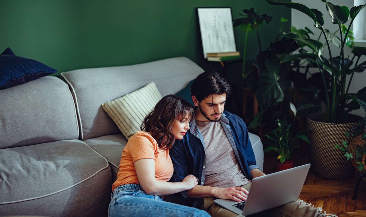 A man and a woman sit on the floor against a couch looking at a laptop