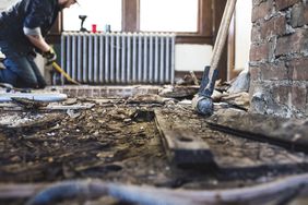 Interior of a home that looks destroyed. Man kneels with a crowbar in the distance. 
