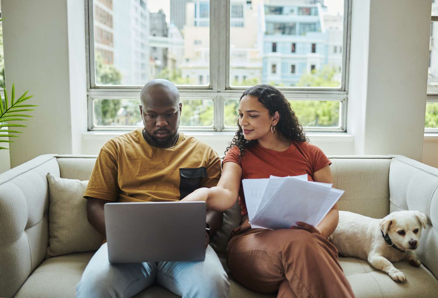 A young interracial couple working on their budget and finances on a sofa from home with their dog using a laptop computer.