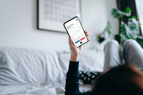 Woman lying on bed at home, reading financial trading data on smartphone