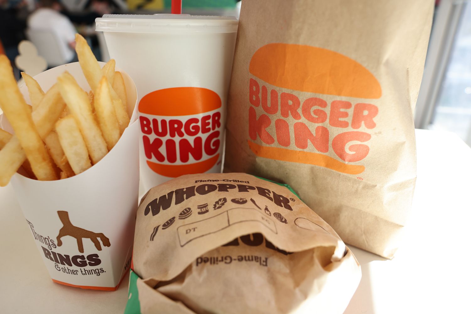 A Burger King bag and drink cup are seen on a table along with French fries and a Whopper cheeseburger.