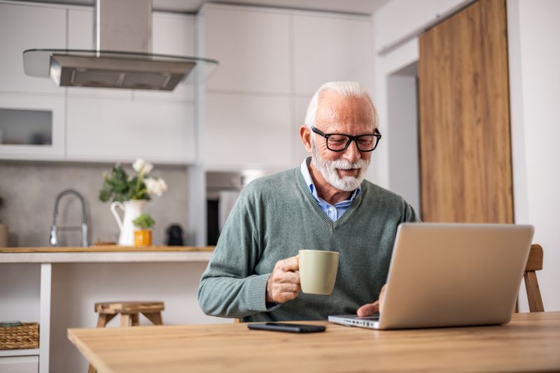 Older homeowner with property in reverse mortgage is looking for information about three-day cancellation rule on laptop while drinking coffee in the kitchen.