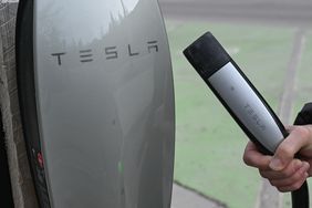 The Tesla logo is seen on the side of a charging station, with a hand holding a charging cable next to the station.