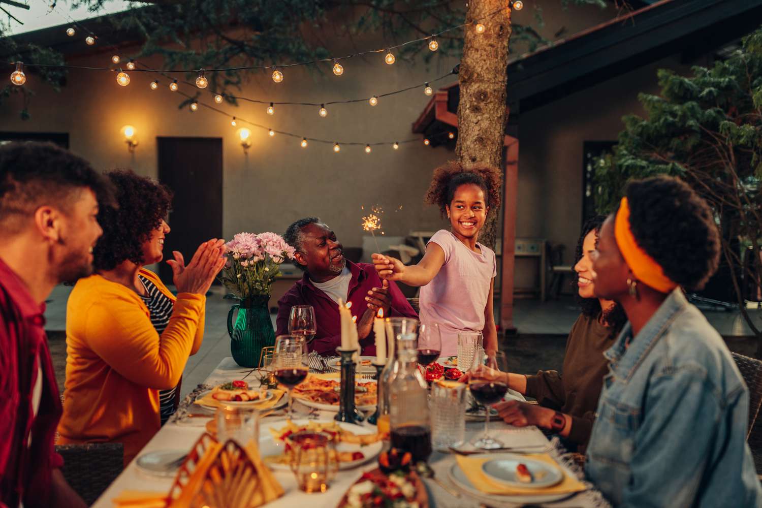 A big family is having dinner in their yard with a happy kid running around the table.