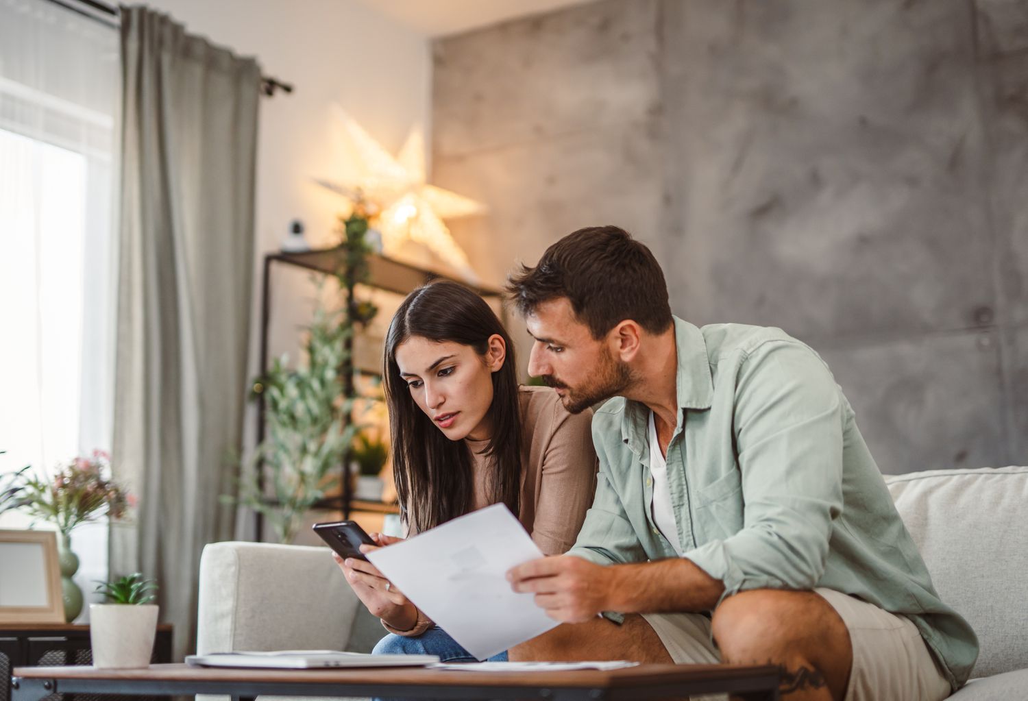 Couple in their 30s sitting on their couch, with holiday decor in the background, looking at documents and a smartphone.